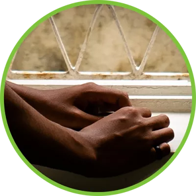Foto mostra as mãos de um adolescente apoiadas no parapeito de uma janela com grades.