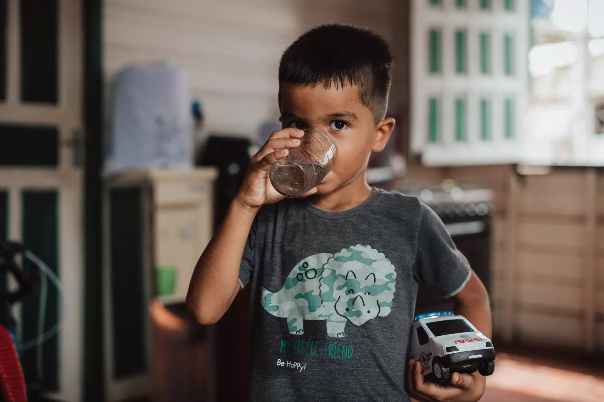 Foto mostra um menino pequeno bebendo um copo de água. Com a outra mão, ele segura um carrinho de brinquedo.Ele está dentro de uma cozinha. 