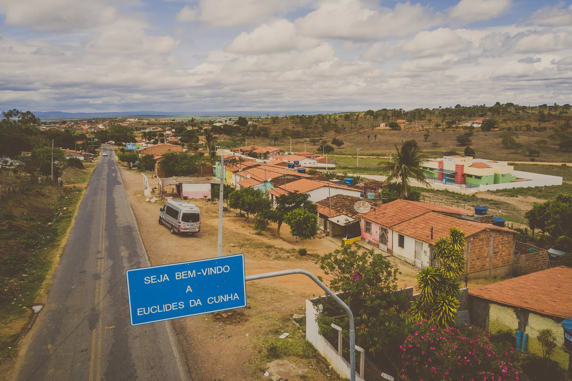 Foto aérea de Euclides da Cunha mostra a placa de limite do município onde está escrito Seja bem-vindo a Euclides da Cunha. Na foto aparecem algumas casas e um ônibus.
