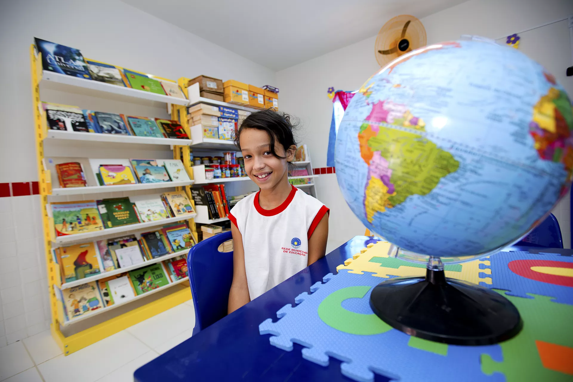 Menina na biblioteca da escola, ao lado dos livros e de um globo terrestre.
