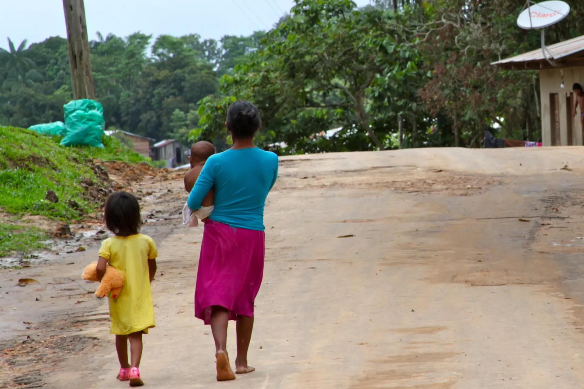 Mulher indígena caminha em estrada de terra com seu bebê no colo e sua filha pequena ao lado