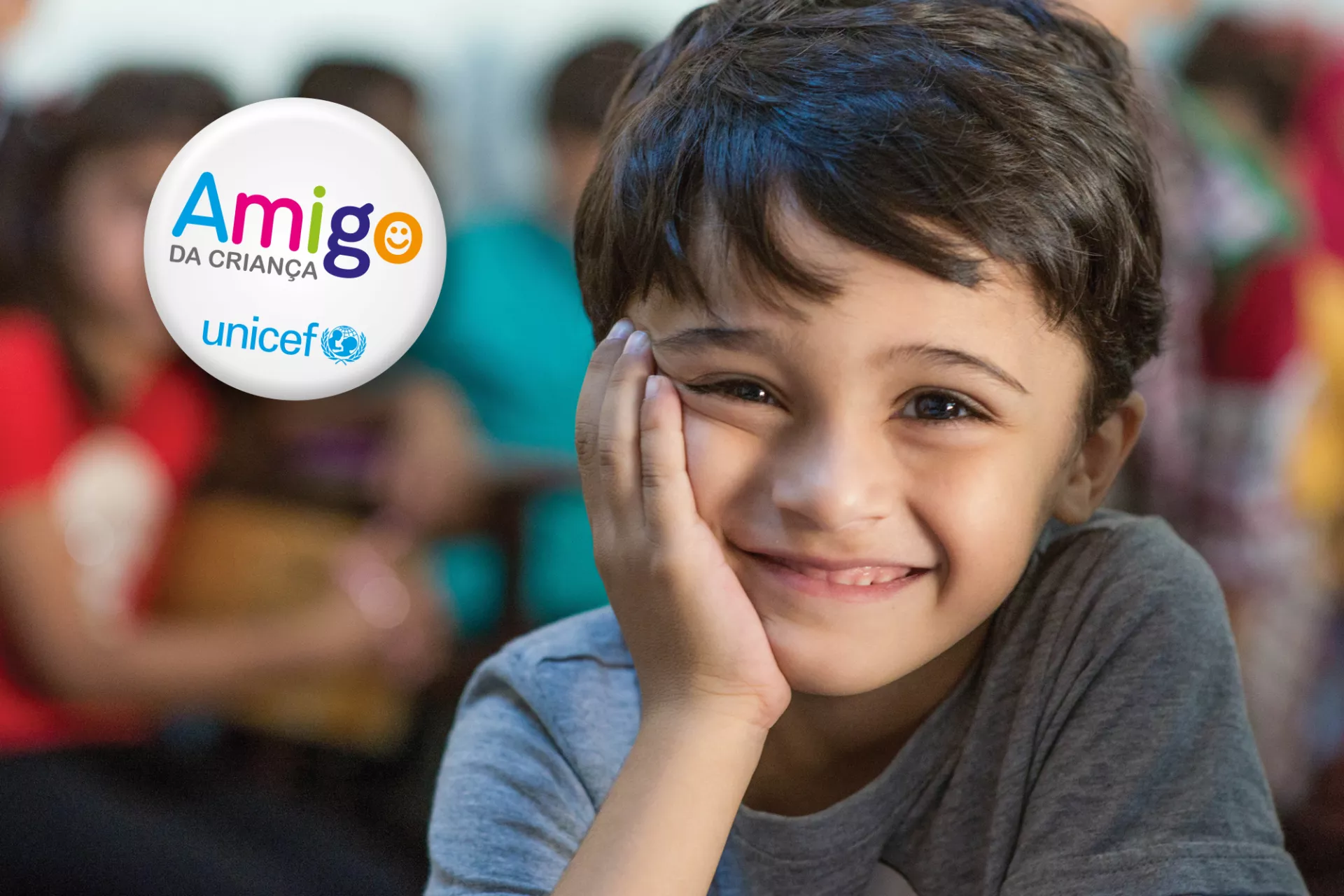 Foto mostra um menino sorridente, apoiando o rosto na mão. Em cima da foto, do lado esquerdo, há a marca do programa Amigo da Criança, do UNICEF Brasil