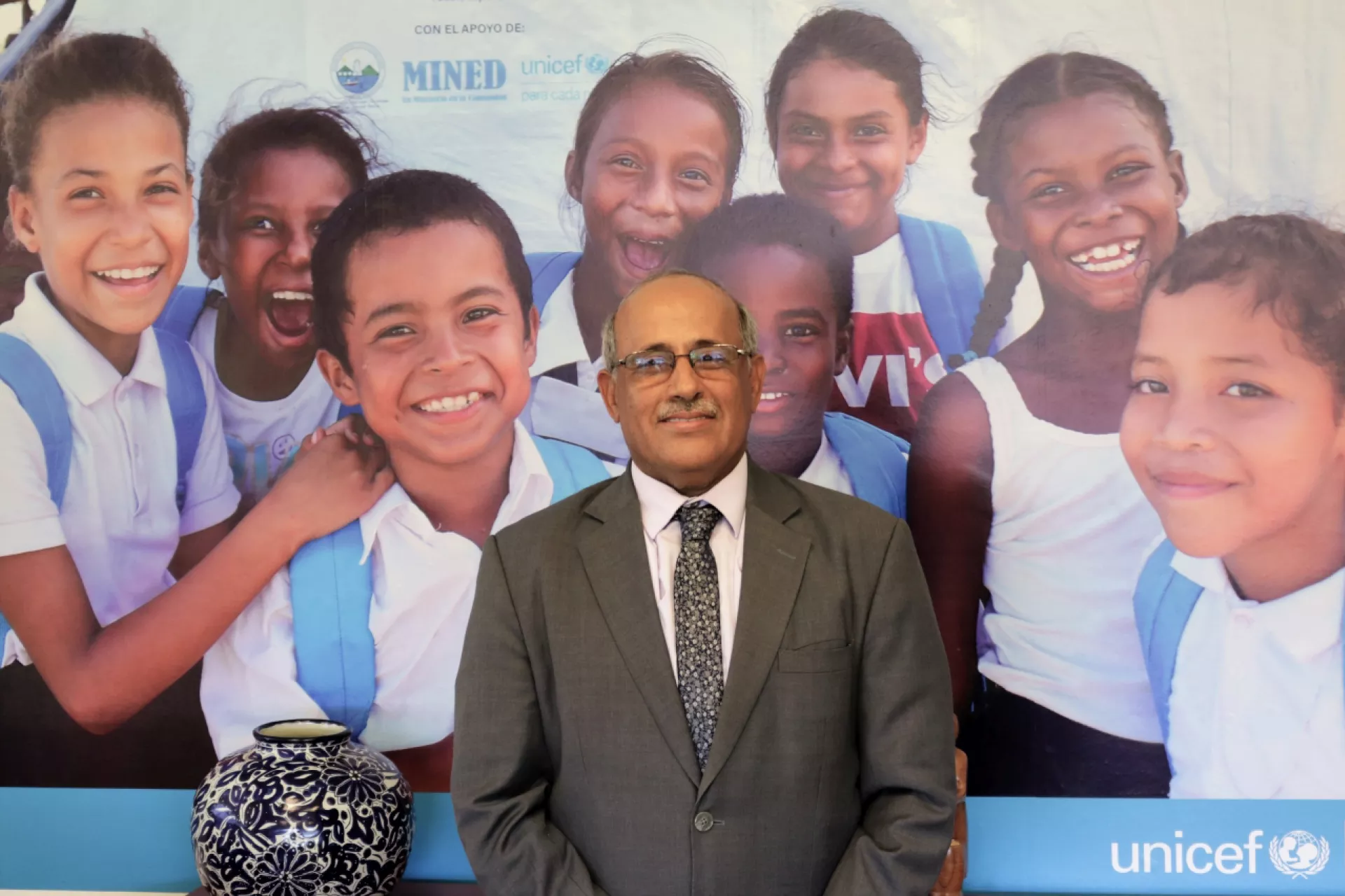 Foto mostra um homem de terno e gravata, olhando para a câmera e sorrindo. Atrás dele há um painel com crianças sorridentes e o logo do UNICEF.