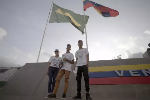 Foto mostra três jovens em pé na frente das bandeiras do Brasil e da Venezuela
