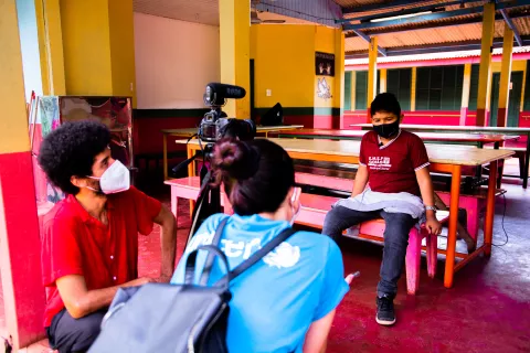 Foto mostra um menino sendo entrevistado por uma funcionária do UNICEF. Na foto aparece também o operador de câmera.