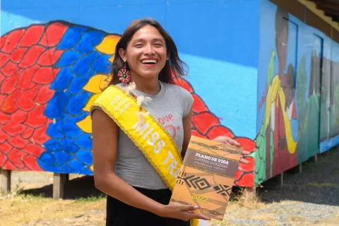 Mulher trans indígena, de cabelos lisos escuros, vestindo uma camiseta de cor cinza e uma faixa amarela, segura um livro e sorri para a câmera. Ao fundo, uma casa com muro azul e desenhos coloridos.