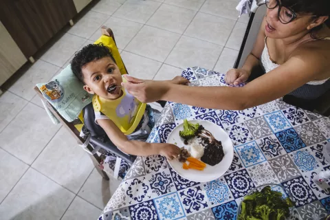 uma mãe dá comida para o filho. a foto está sendo tirada de cima e o menino levanta a cabeça para olhar para a câmera