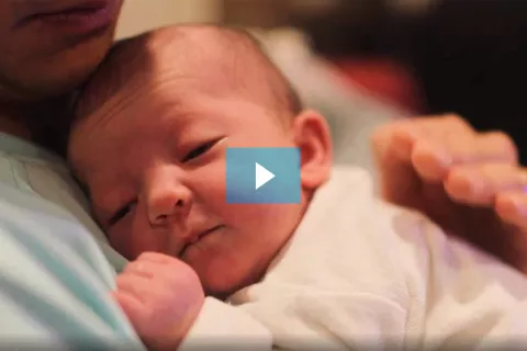 Foto extraída do vídeo mostra um bebê no colo do pai. O pai está batendo com a mão nas costas do bebê para que ele arrote.