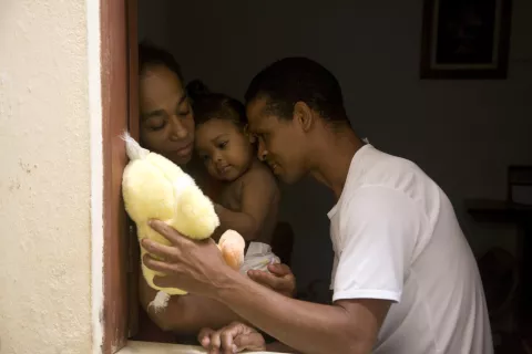 um pai mostra um ursinho de pelúcia amarelo para sua filha bebê que está no colo da mãe. a cena é toda muito carinhosa.