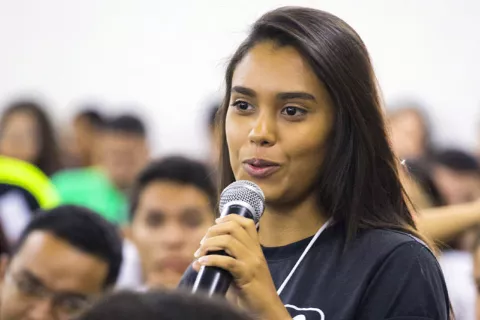 Foto mostra uma adolescente falando para outros adolescentes e jovens. Ela está segurando um microfone