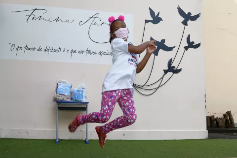 Uma menina salta com as pernas para tras em frente a uma parede onde estão desenhados pássaros.