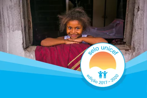 uma menina está olhando pela janela da casa dela. em cima da foto há uma onda azul e o logo do Selo UNICEF