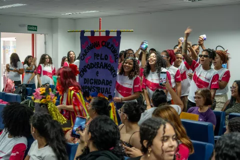 Adolescentes reunidos em uma sala, uma adolescente segura um estandarte onde se lê Caravana Cultural Pelo Empoderamento e Dignidade Menstrual