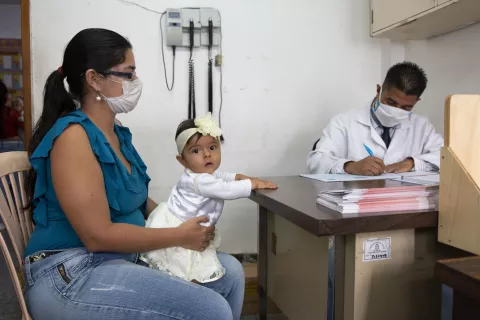 uma mãe está em um consultório médico com sua bebê. ela e o médico usam máscara. a bebê está olhando para a câmera