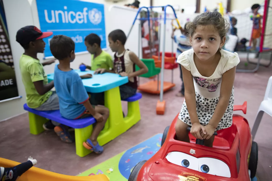 menina brinca com um carrinho em um abrigo para crianças venezuelanas em Roraima. Ao fundo, outras crianças brincam em uma mesa.