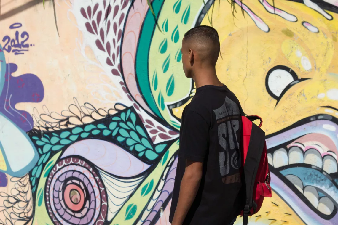 Adolescente de costas, carregando uma mochila, passa em frente a um grafite de rua