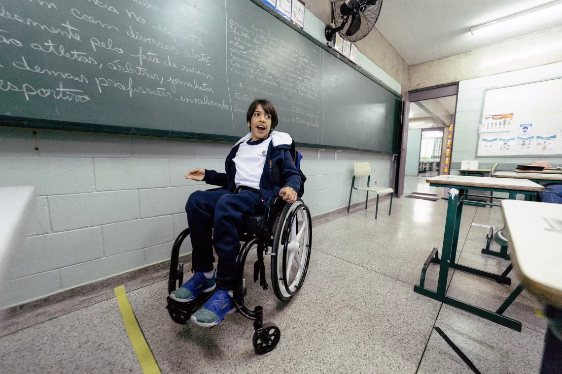 Foto mostra um adolescente em uma cadeira de rodas em frente a um quadro-negro dentro de uma sala de aula