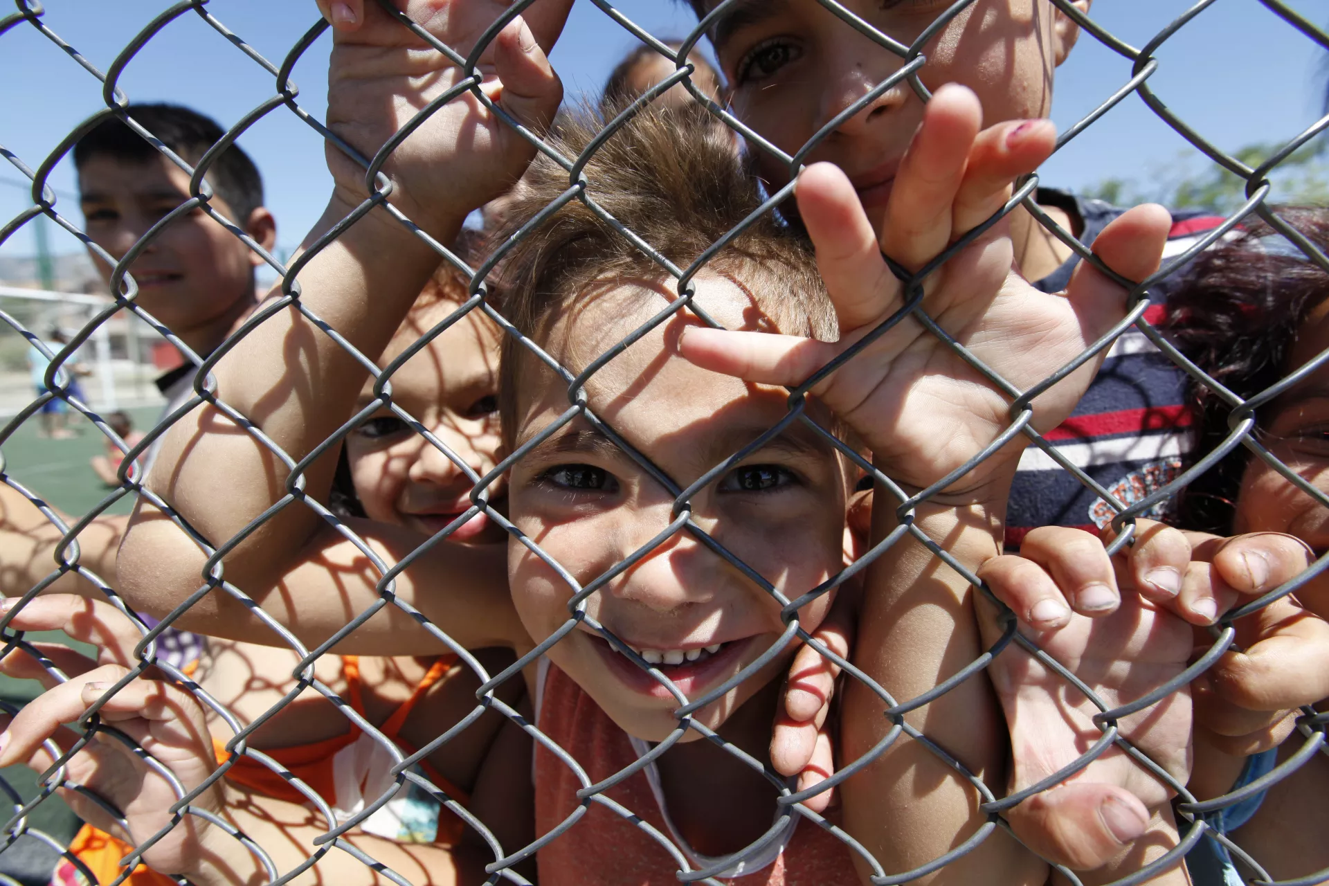 Деца позират усмихнати, скупчени зад оградата на спортна площадка.