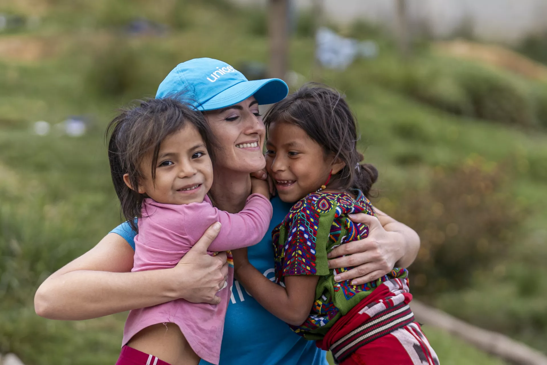 Woman wearing UNICEF hat and t-shirt hugs two young girls. They all smile.