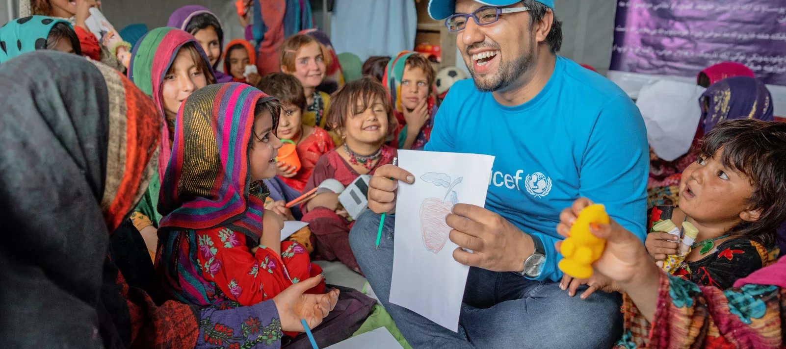  A man wearing UNICEF branded t-shirt sitting on the ground amid a group of children in a classroom in a school in Afghanistan.