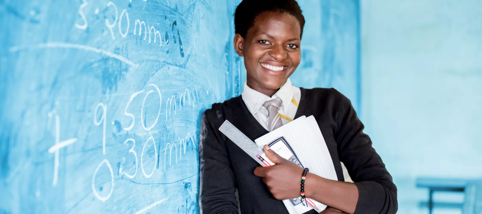A smiling adolescent girl, holding school supplies and a calculator, leans against the blackboard during a class, in Kamulanga Secondary School in Lusaka, the capital.