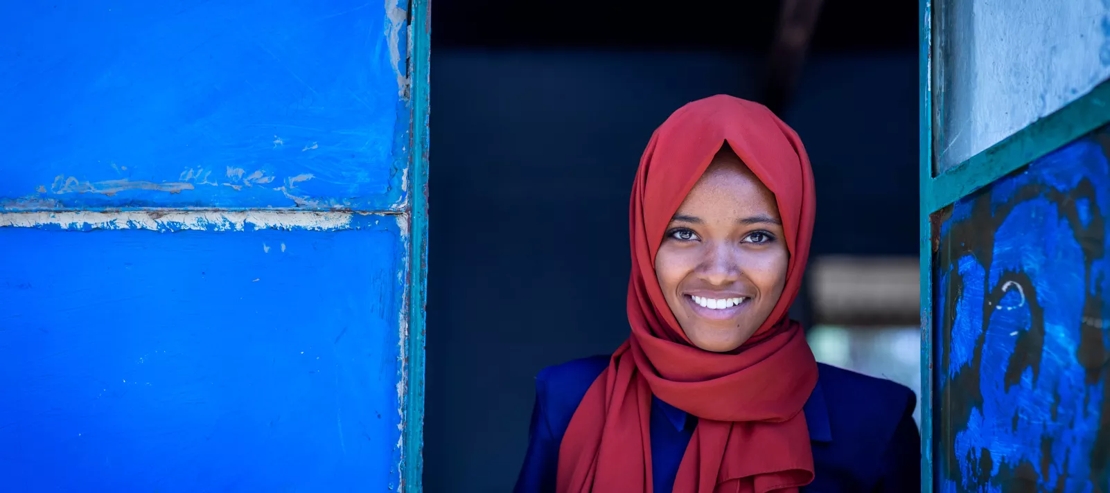 A girl in an orange headscarf smiles as she leans out of a window