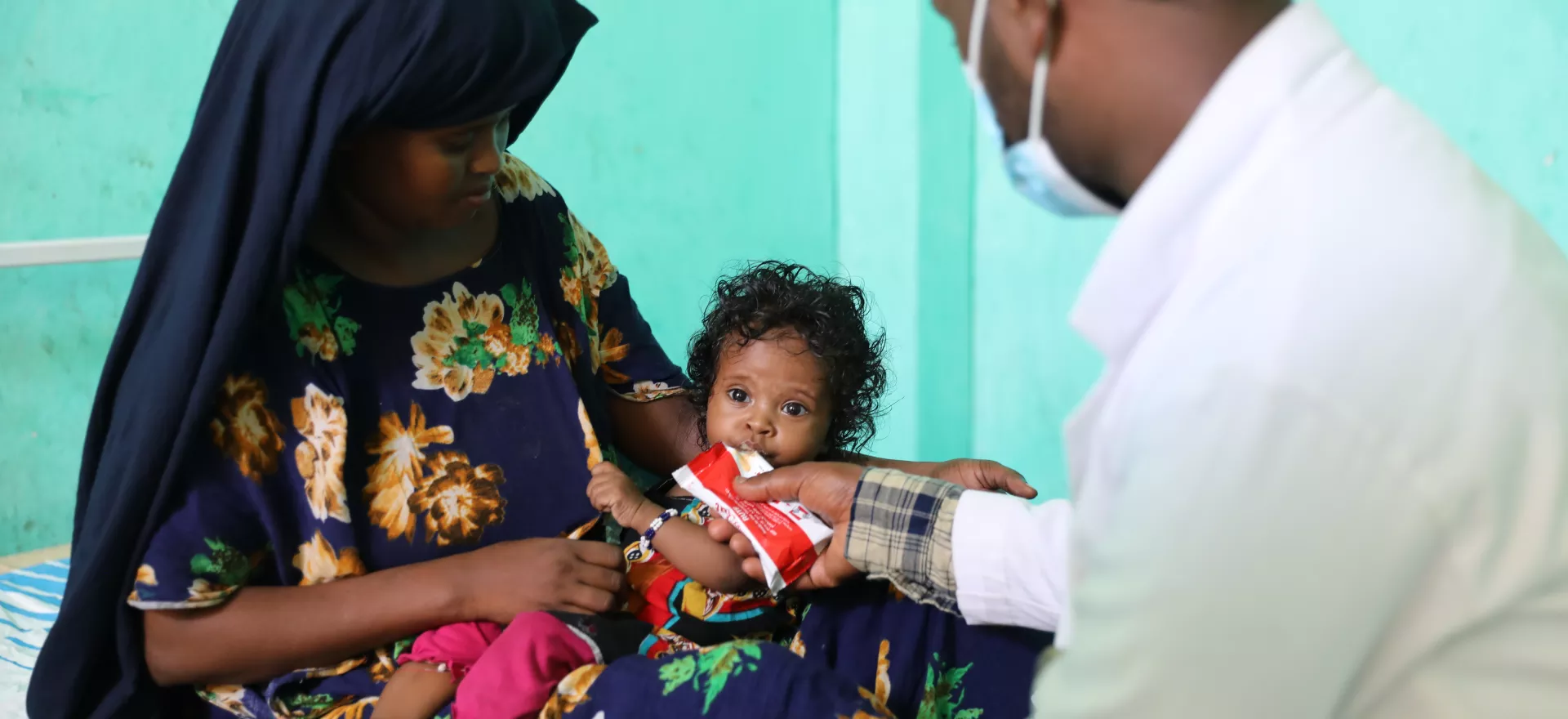 A health worker holds a sachet of therapeutic food for a little girl, held by her mother to eat.