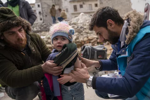 Male health team worker looks at a toddler's arm with rubble from a collapsed building in the background.