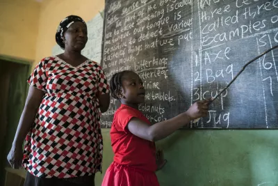 A teacher supervises a schoolgirl reading from the blackboard