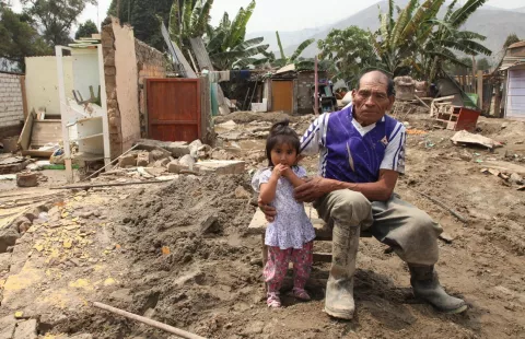 Un adulto mayor y una nina sentados frente a una casa destruida por un aluvión