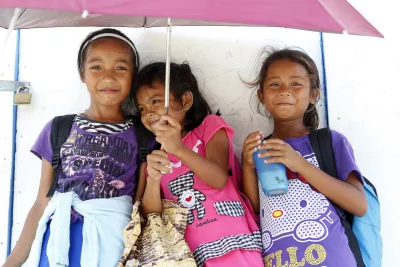Three children carrying school bags gather under an umbrella, smiling