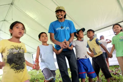 A UNICEF staff member, wearing a blue UNICEF t-shirt with the URL for the UNICEF Philippines website, holds hands with children participating in a recreational activity inside a UNICEF tent.