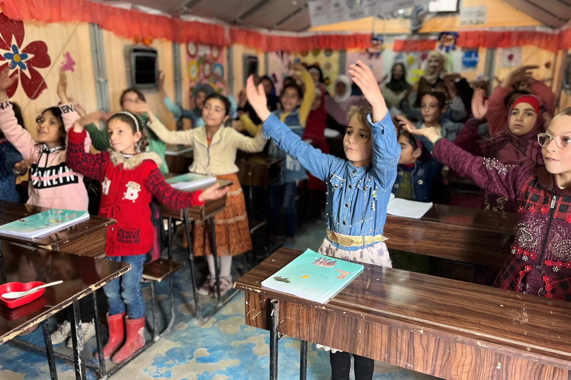 Syria. A group of children raise their arms as they sing along in a classroom.