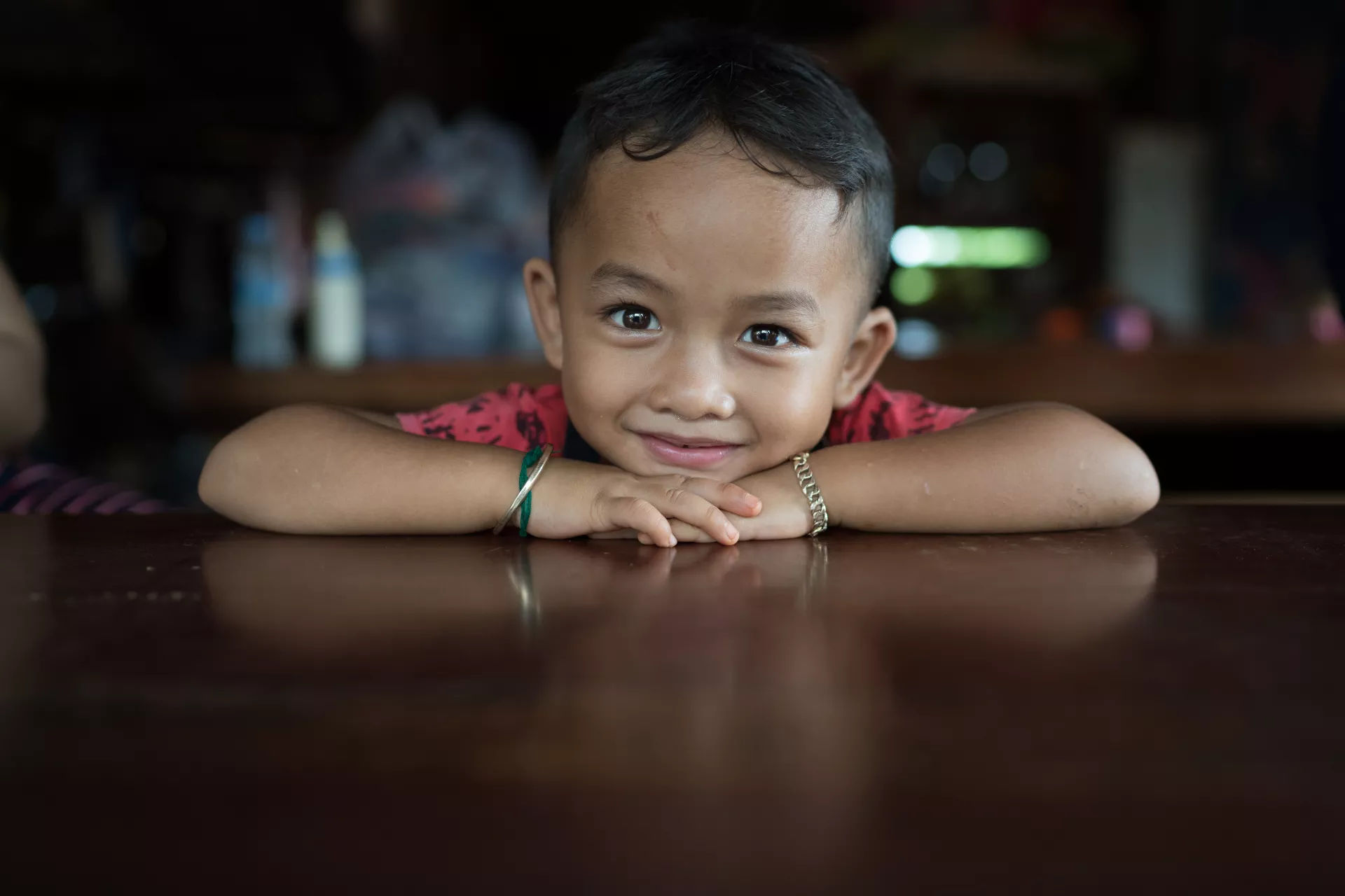 A four-year-old boy rests his head on his hands at a table in his house, smiling.