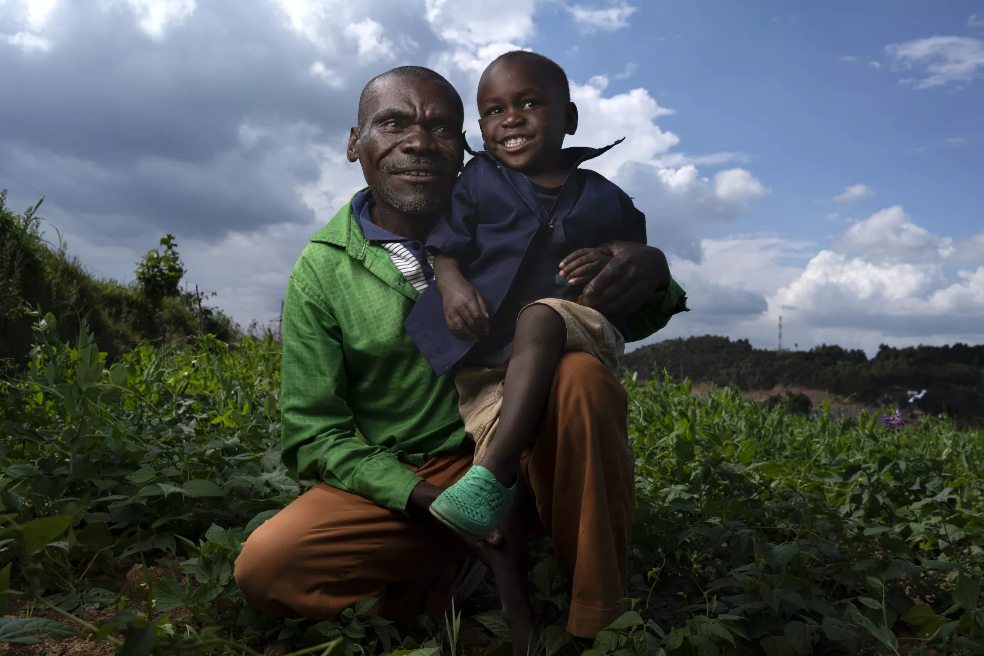 A father squats with his two-year-old son in his lap, both smiling, in a field in Rwanda.