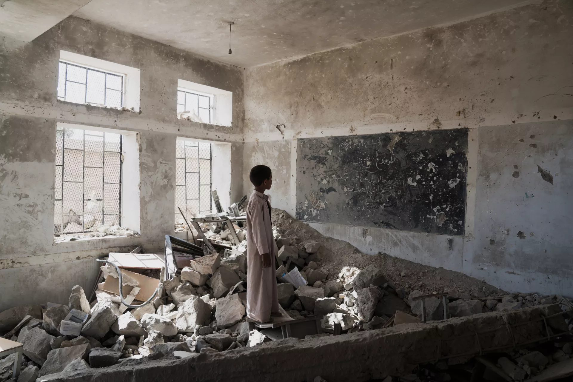 A young child stands in a classroom of rubble in Saada, Yemen, staring at what used to be a chalkboard.