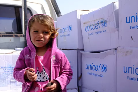 Une petite fille portant un pull à capuche rose, se trouve près de cartons blancs avec écrit UNICEF dessus