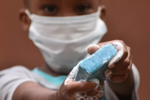 A boy wearing a face mask holds out a bar of blue soap in his hands