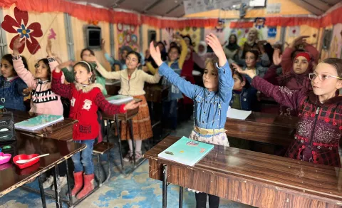 Syria. A group of children raise their arms as they sing along in a classroom.
