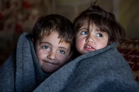 Deux enfants serrés l’un contre l’autre, emmitouflés dans une épaisse couverture grise, sous la tente où vit leur famille dans un camp de réfugiés en Iraq.