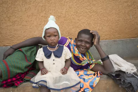 A mother lies on her side, head in hand, with her baby seated in front of her outside a health centre in Uganda.