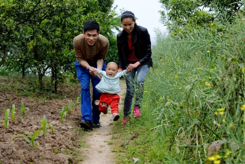 Two parents walking down a dirt path near the woods play with their young son.