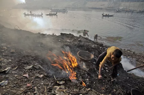 Standing on a mound of debris, a boy burns garbage to expose discarded scrap metal, on the banks of the Buriganga River, in Hazaribagh, Bangladesh.
