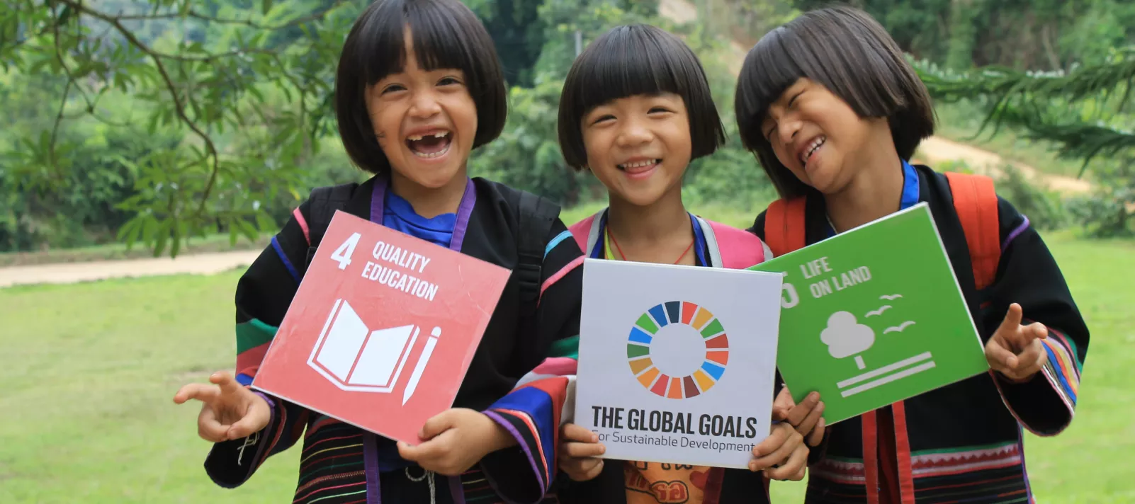Three young girls smile outside in Thailand while posing with posters for the SDGs.