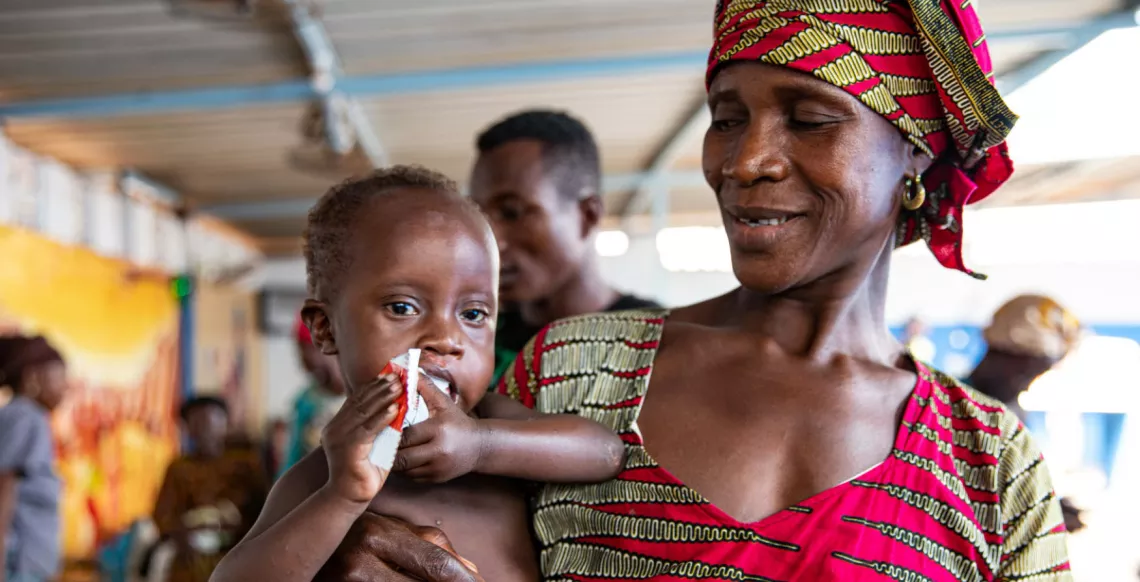 A woman holds a child who is eating from a package of therapeutic food