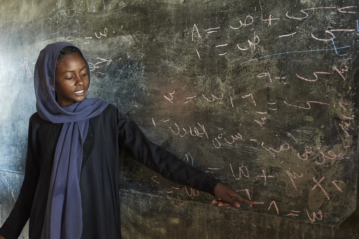 A young girl stands in front of a chalkboard facing her class to explain a math equation.