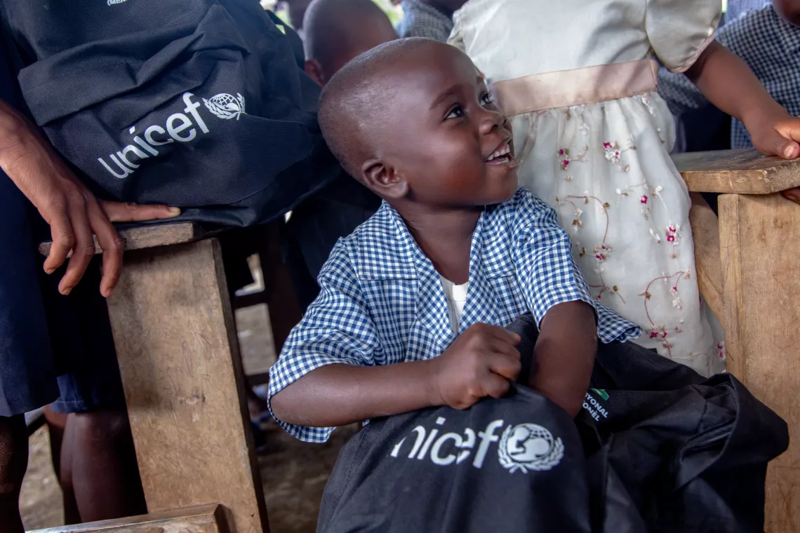 A boy smiling brightly sticks his hand into a black school bag with the UNICEF logo that he is holding in his lap.