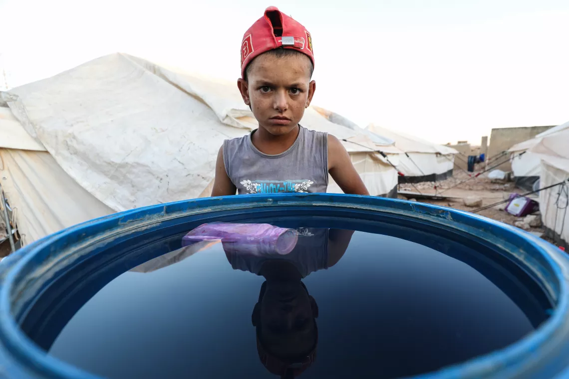 Syria. A small boy stands nexts to a water container.