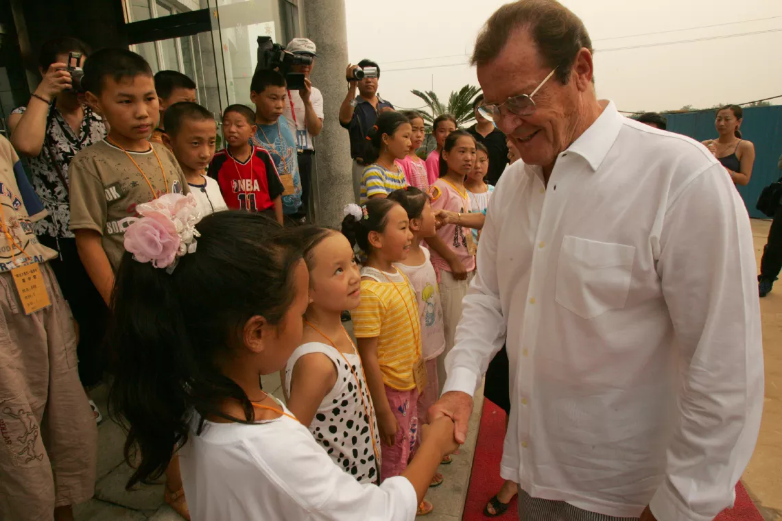 On 10 August 2004 in China, UNICEF Goodwill Ambassador Sir Roger Moore (right) greets a girl, as other children and members of the press look on, during the launch of a UNICEF-supported summer camp for children orphaned by AIDS, in Beijing, the capital.