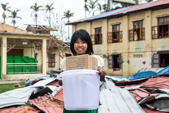 Myanmar: A girl returning from school in school uniform receiving a water bucket and hygiene kit, including laundry and body soaps, and sanitary pads distributed by UNICEF.